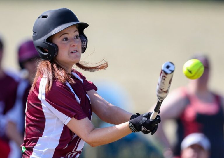 Close-up action shot of a female player in maroon kit making contact with a softball, ball visible near the bat