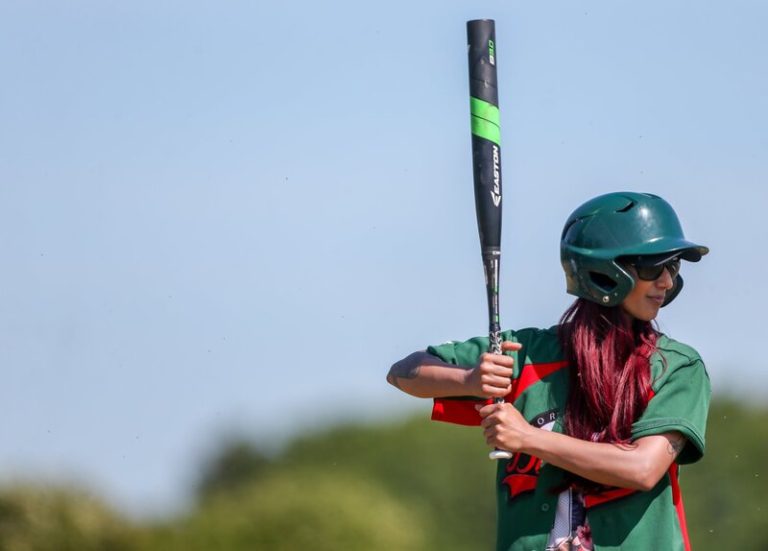 Player in green and red kit in a ready batting stance with helmet on and bat raised, bright blue sky and green field behind them