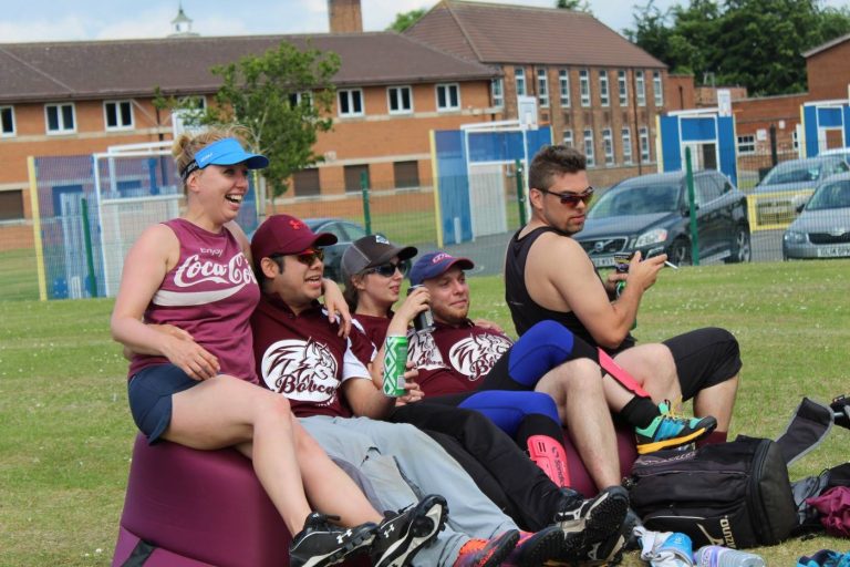 Group of Bobcats players in maroon kit sitting and watching play from the sideline on a sunny day, kit bags and equipment beside them