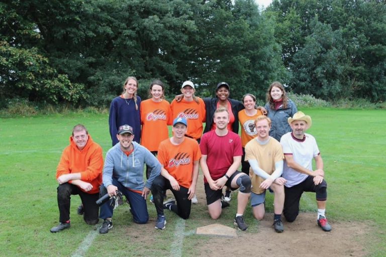 Mixed group photo including Coyotes players in orange and players from other teams in various kits, on a softball field with trees behind