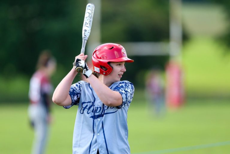 Hazel in batting stance wearing a blue kit and red helmet, bat raised ready to swing at an outdoor softball field