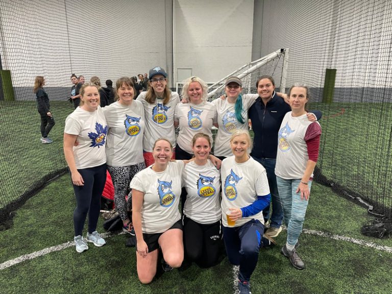 Leeds softball team group photo inside a sports hall, women wearing white Leeds-branded shirts, posed in front of goal netting