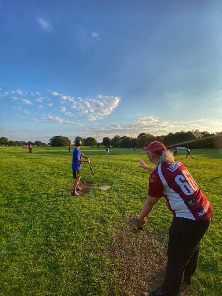Wide field action shot with a player in red and white near a base, other players visible in the background on a grass field
