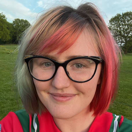 Headshot portrait of Chuck in a green and red Dales kit, smiling outdoors with pink and red hair
