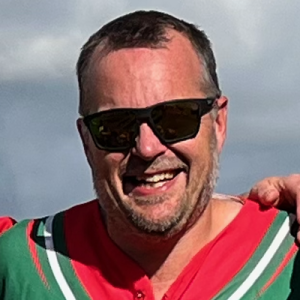 Close portrait of Dales player Glover in sunglasses and red and green kit, smiling in bright sunshine on the field