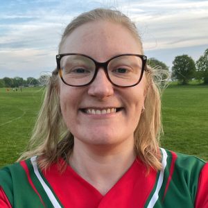Headshot portrait of Pippa in a green and red Dales kit with glasses, smiling on an outdoor field