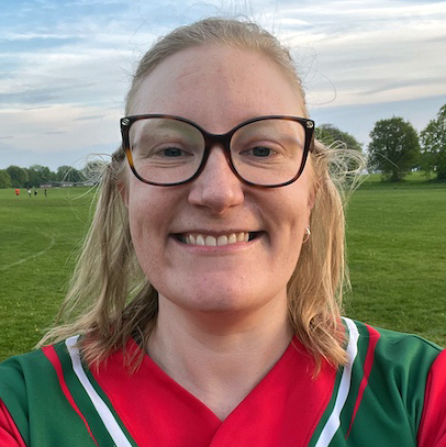 Headshot portrait of Pippa in a green and red Dales kit with glasses, smiling on an outdoor field