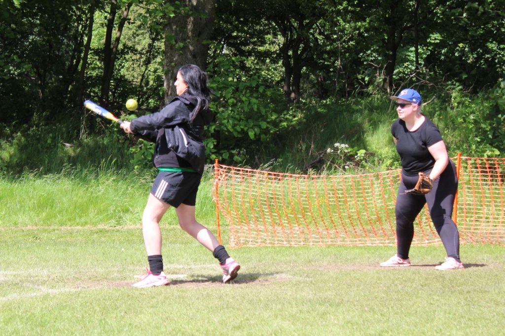 Player in green kit taking a swing at a batting net, ball in the hitting zone, practice netting and treeline visible behind