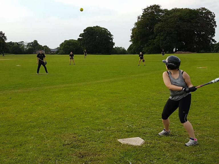 Player in dark kit fielding in the outfield on a bright green pitch, ball visible in the air above and other players spread across the field