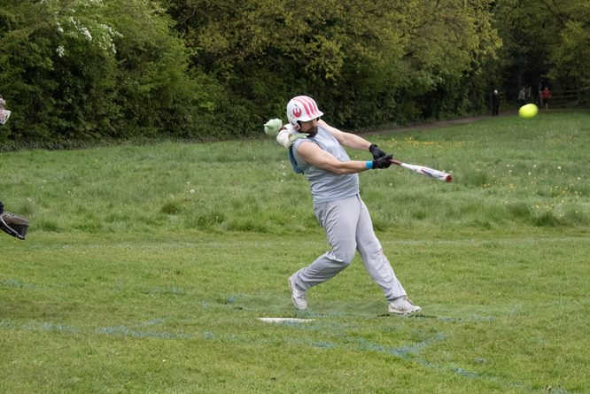 Batting Action Grey Kit Softball player in grey kit driving through a full batting swing on a grass field
