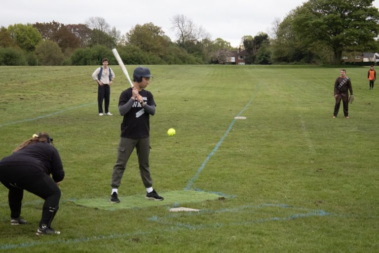 Player in dark kit taking a swing at a batting tee, ball in the hitting zone, open green field around them