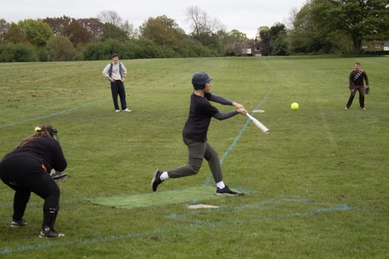 Player in dark kit fully extended through a batting swing at the tee, coach observing from behind on the practice field