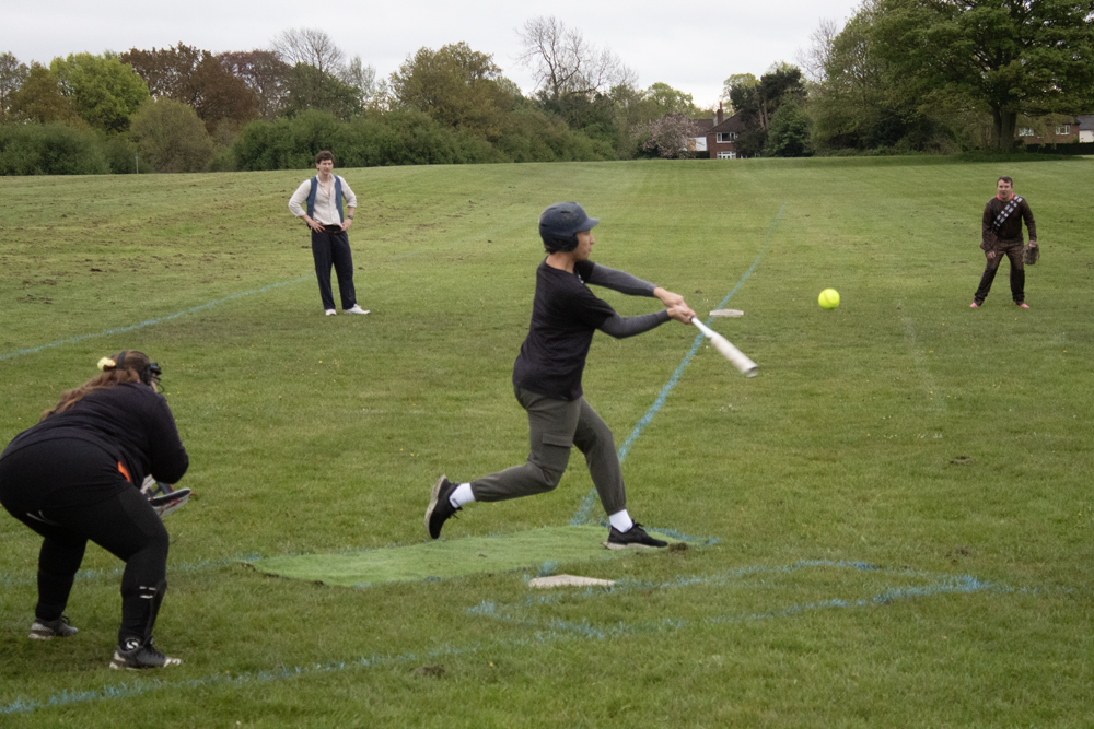 Batting Tee Full Extension Player in dark kit fully extended through a batting swing at the tee, coach observing from behind on the practice field