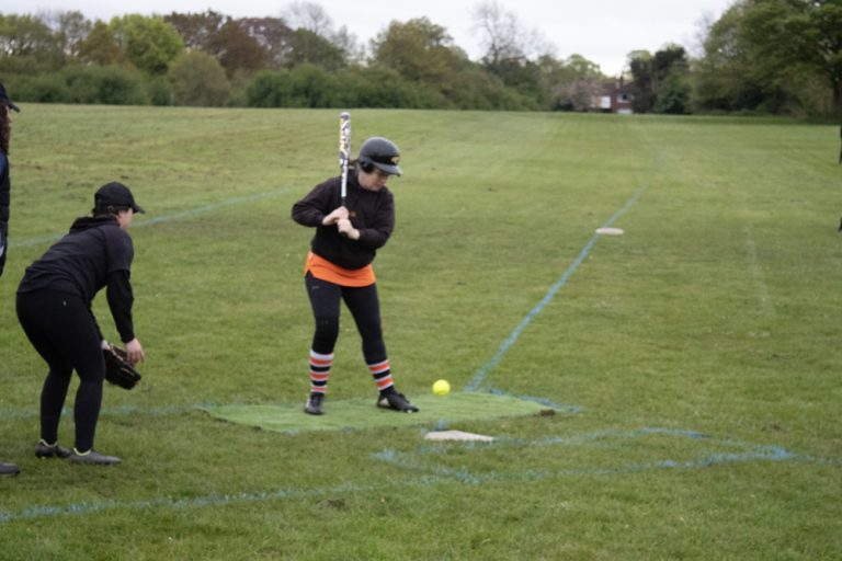 Player in orange and black kit at a batting tee station preparing to swing, coaches and other players watching nearby