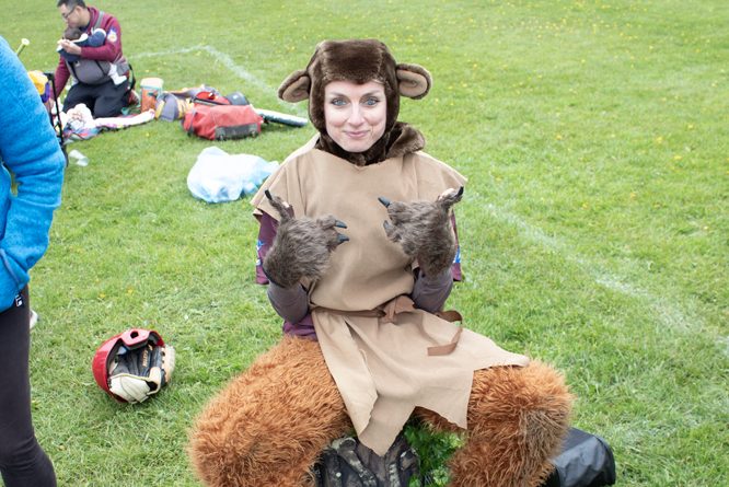 Bear Costume Sideline Person in a brown bear costume sitting on the grass at the edge of a softball field