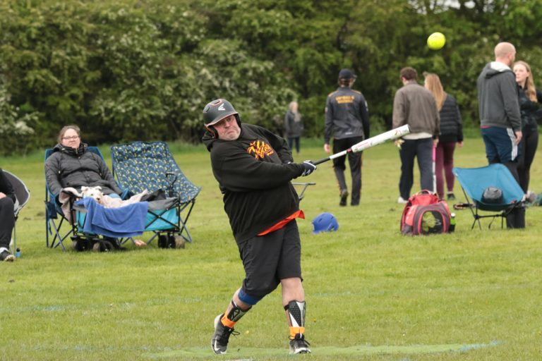 Player in black kit completing a powerful full batting swing, ball in frame, spectators and kit bags visible in the background
