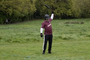 Softball player in maroon uniform completing a bat swing follow-through on a grass field