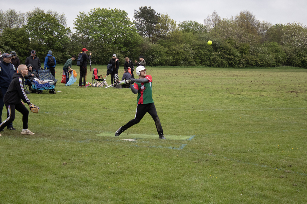 Green Kit Batting Swing Player in green kit completing a batting swing at a tee station with good body rotation visible, overcast sky above