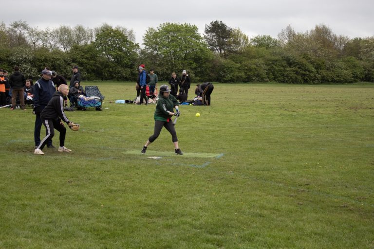Player in green kit at a batting tee mid-swing, other players and spectators gathered near the boundary watching on