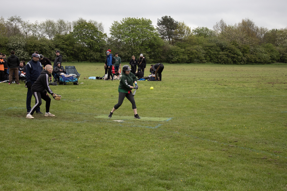 Green Kit Batting Tee Player in green kit at a batting tee mid-swing, other players and spectators gathered near the boundary watching on