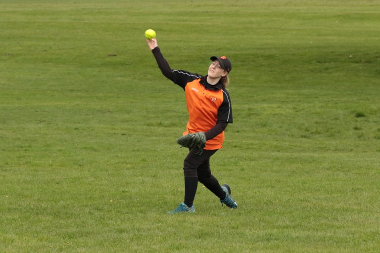 Player in an orange training bib reaching up to catch a softball in the field, arms outstretched on a green pitch