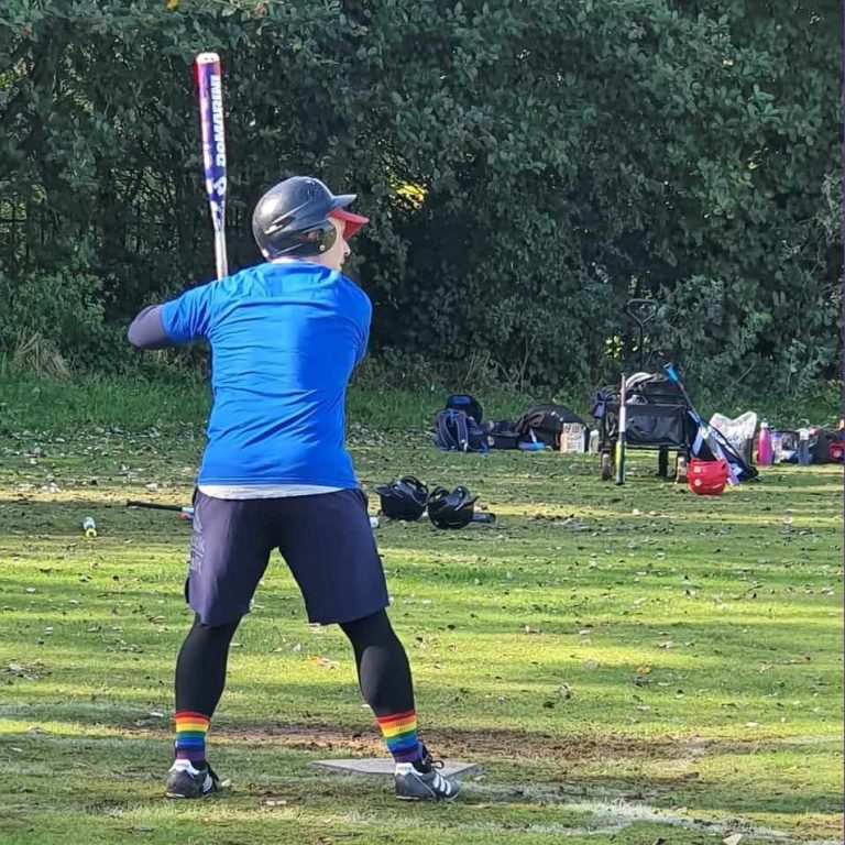 Player in blue kit and helmet in a full batting swing, bat extended, trees and greenery visible in the background