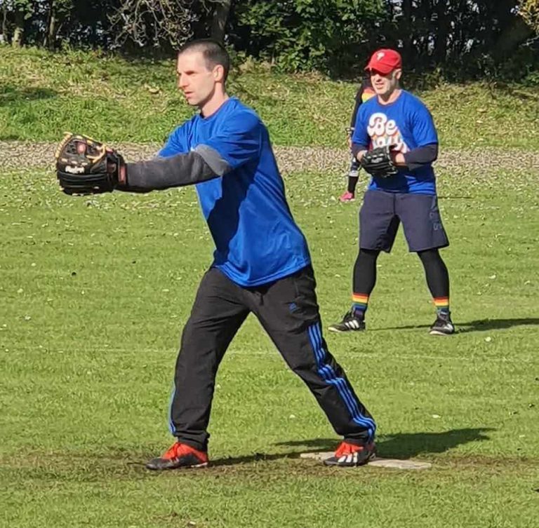 Player in blue kit and glove fielding a ground ball in the infield, second player visible in the background on a sunny day