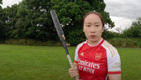 Young female softball player in an Arsenal FC shirt holding a softball bat vertically, standing on a green field