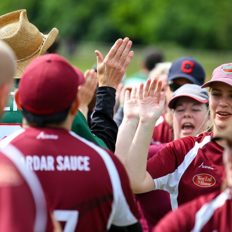 Softball players in maroon kit forming a high-five line during a sunny match day, smiling and celebrating