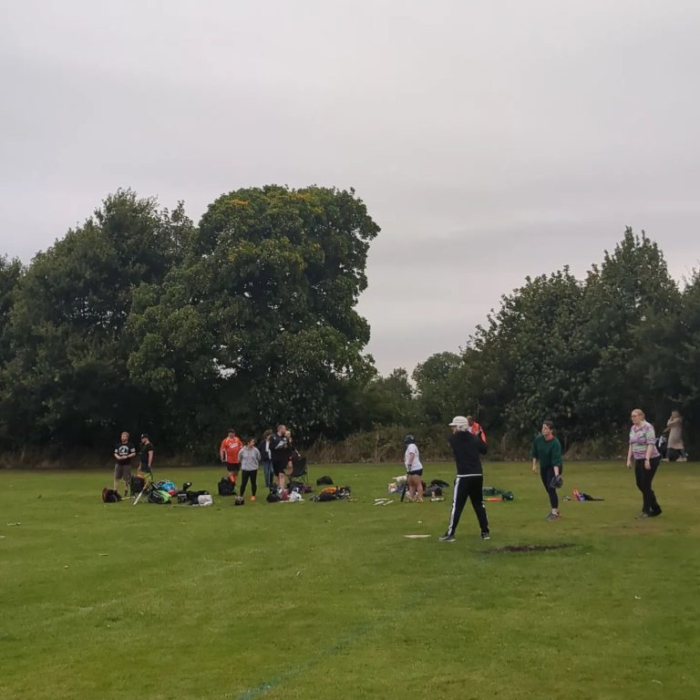 Group of softball players warming up and gathering on a green field under a grey overcast sky, trees visible in the background
