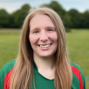 Headshot portrait of Lucy in a green and red Dales kit, smiling outdoors with trees in background