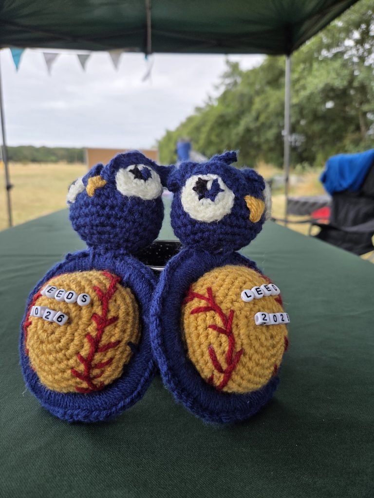 Two hand-crocheted softball mascots, blue faces stitched onto yellow balls, on a table under the tournament gazebo